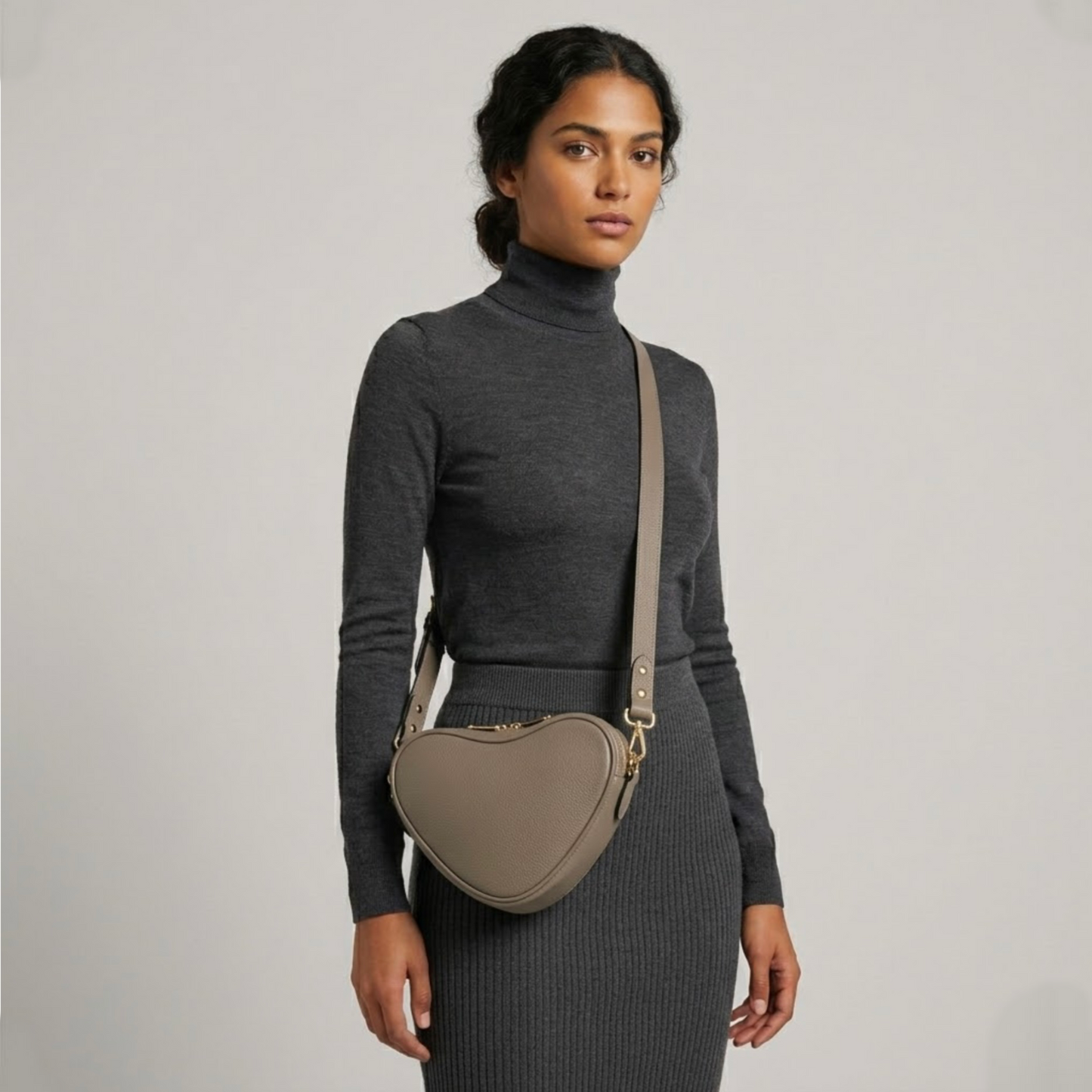 Beige heart-shaped leather crossbody bag by Tikori London, featuring a stylish and compact design. Beautifully dark skinned model wear the bag as a crossbody. She wears grey top and grey tight skirt . Bags long strap and shimmering golden hardware shows in a soft grey background.