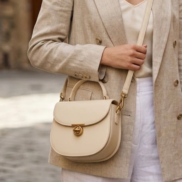 Luna Saddle Bag - Ivory by TIKORI LONDON. The centrepiece is a custom engineered gold T-bar turn lock, providing a secure closure and a sophisticated focal point. Body of a model showing wearing the bag as a crossbody in Mediterranean background. She wears white cotton trousers and cream blazer. Bag featuring Golden tone hardware and long strap. Front profile of the bag in the picture.