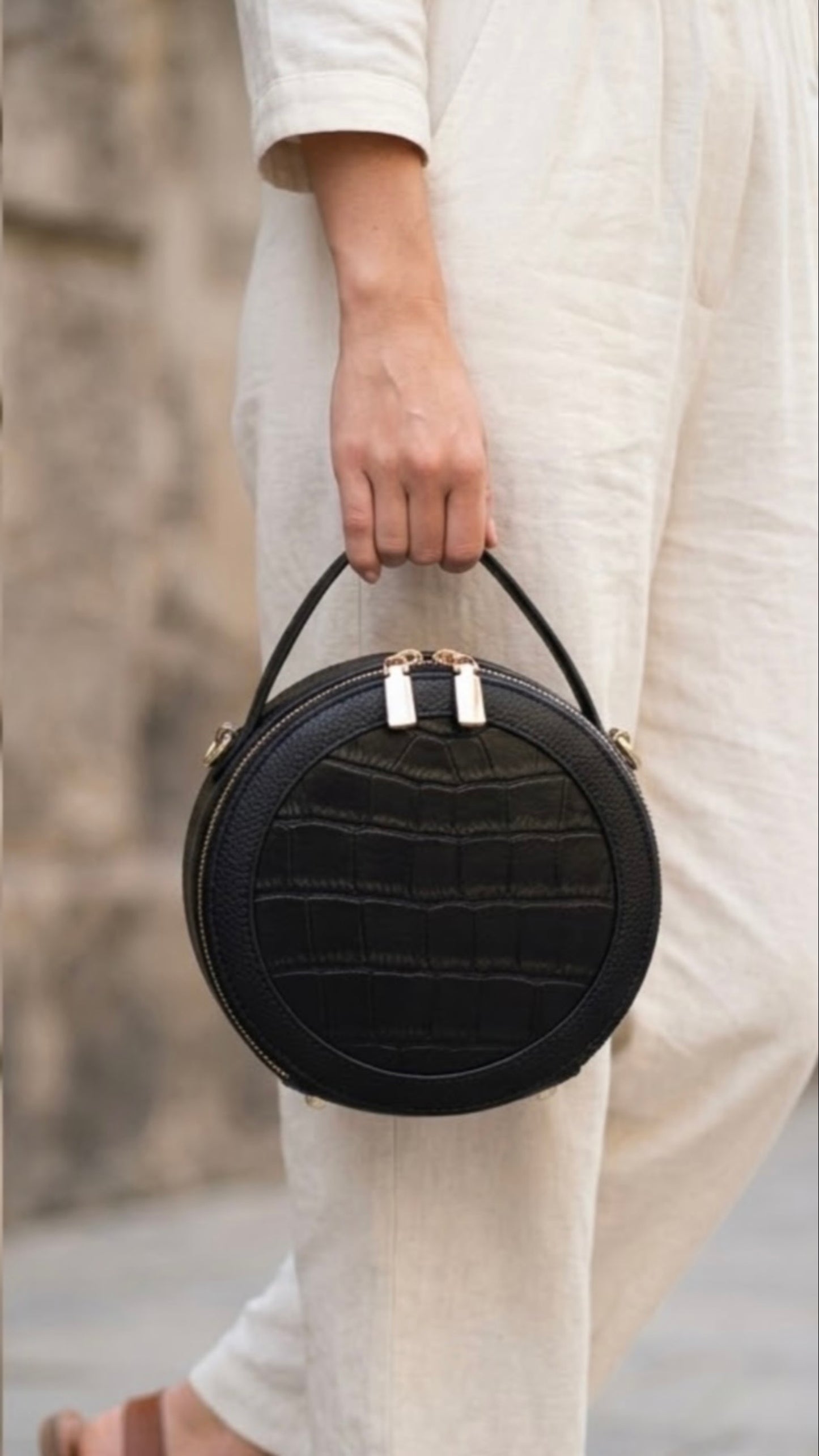TIKORI LONDON black finished leather circle crossbody bag with dual zip fastenings, featuring a sleek, rounded design. Body of a model in ivory cloths carrying the bag from top handle. Front profile of the bag with golden hardware shows in the picture