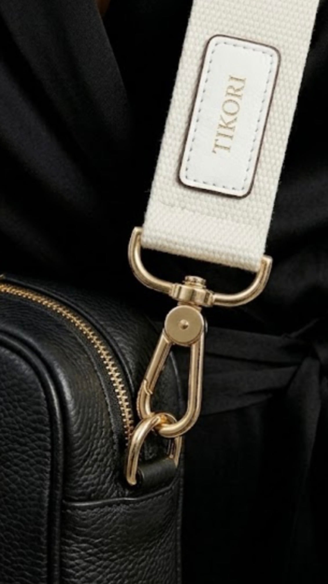 Classic elegance with practicality and versatility in mind. TIKORI classic Black by TIKORI london. Model in black cloths, wears the crossbody. Close up detail of the canvas strap and golden tone hardware shows in the picture with TIKORI logo on the canvas strap.
