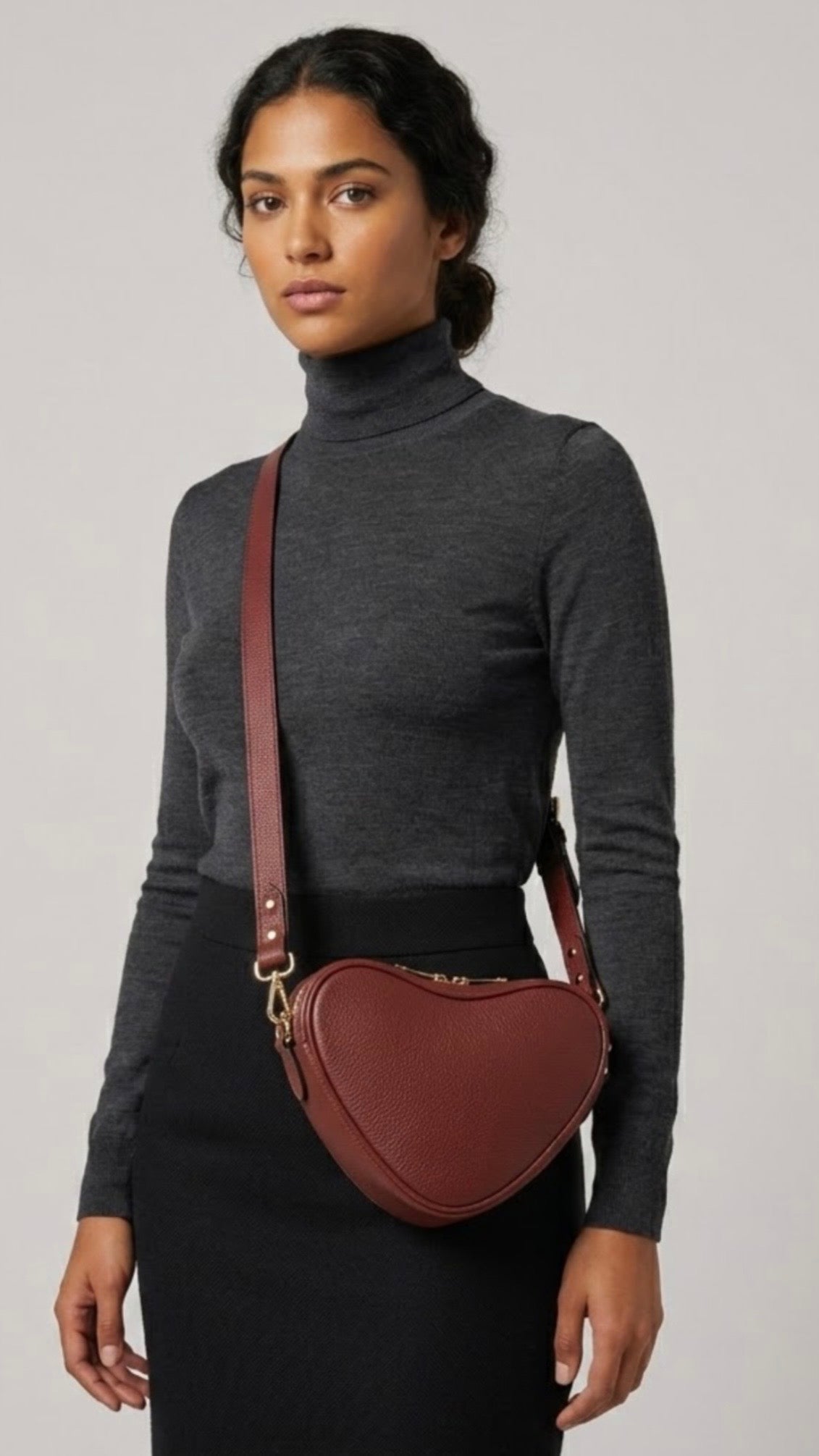 Red brown heart-shaped leather crossbody bag by Tikori London with adjustable strap and gold-tone hardware. Dark skinned beautiful model in black tight skirt and dark grey top wears the crossbody. Front profile of the bag showing in the picture with golden tone hardware and long strap highlighted in soft creamy background.