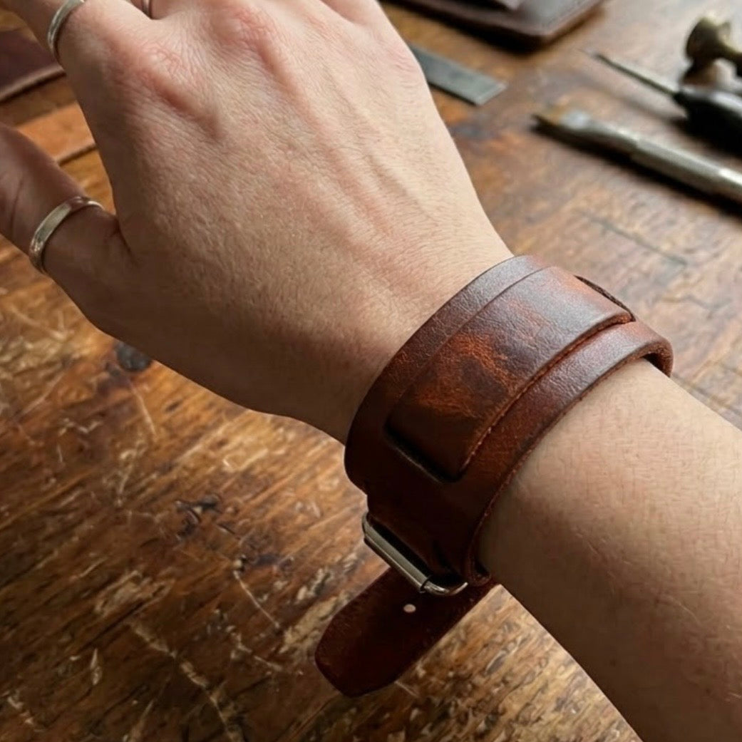 Dark brown leather layered bracelet by TIKORI London with smooth leather layered look, stylish accessory for men and women. Male model hand displays wearing the dark brown bracelet. Table with leather items and tools also shows in the picture. Leather workshop environment.