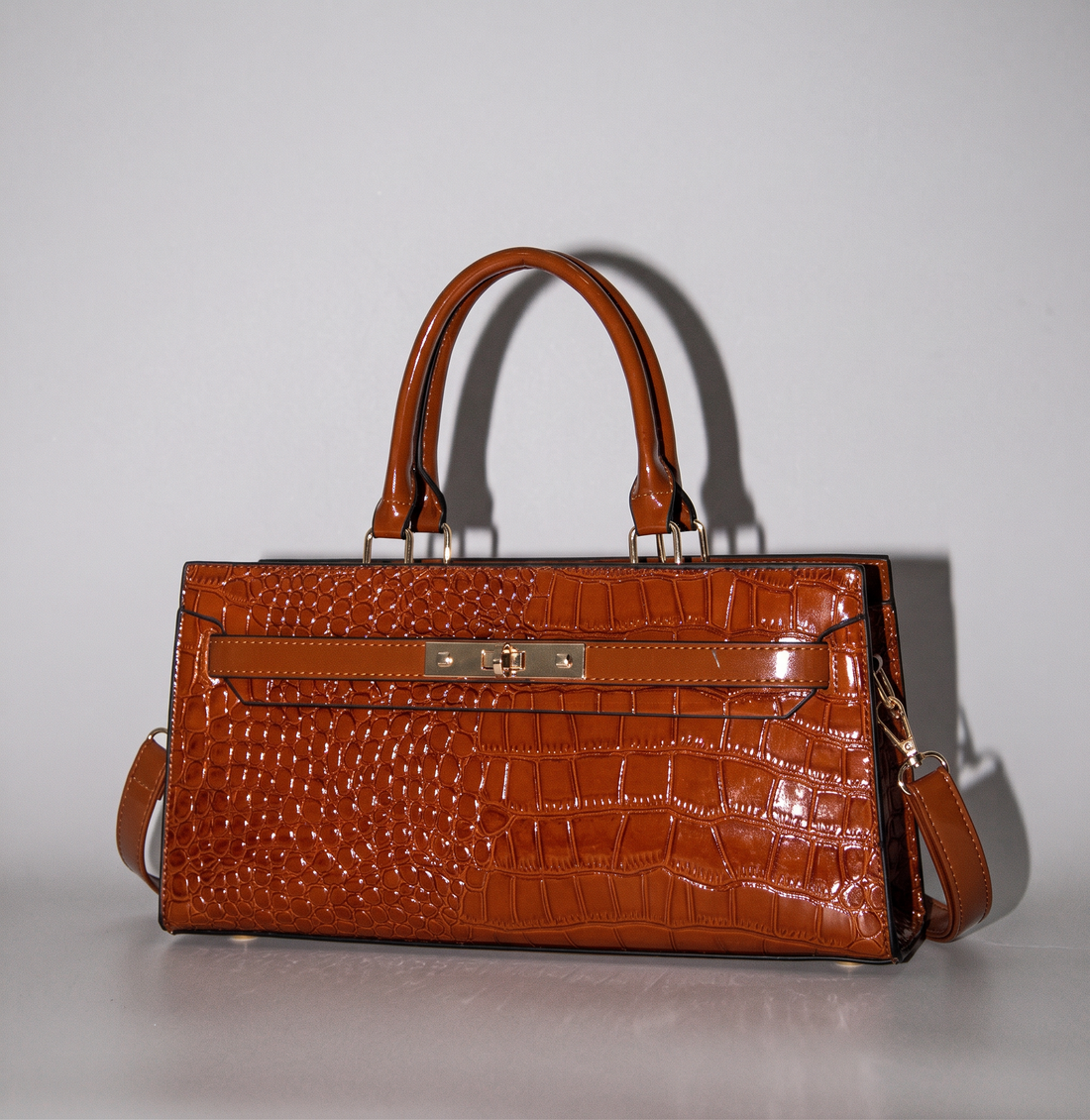 The Gilded Croc Baguette Shoulder Bag by TIKORI LONDON. beautiful croc finished vegan leather bag with top handles displayed in the picture. Front golden fasteners shows in a fully light grey background. Long strap also attached to the bag. 