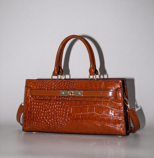 The Gilded Croc Baguette Shoulder Bag by TIKORI LONDON. beautiful croc finished vegan leather bag with top handles displayed in the picture. Front golden fasteners shows in a fully light grey background. Long strap also attached to the bag. 