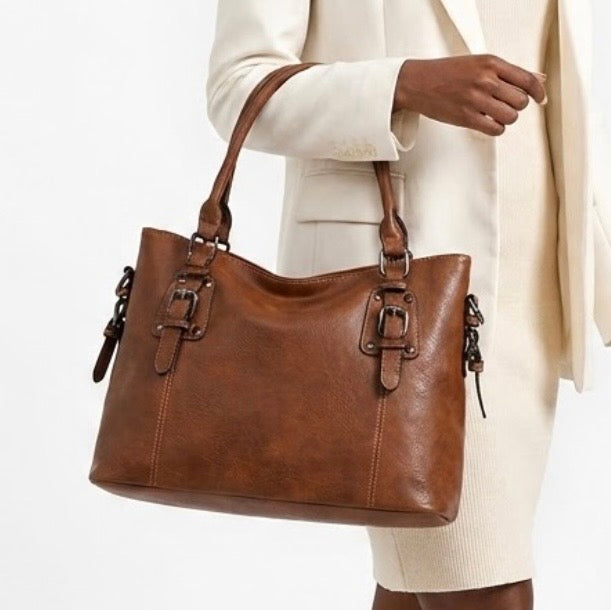 Maven  Satchel - Warm Brown by TIKORI LONDON. Constructed from deep-brown, pebbled Vintage washed Faux Leather with a supple hand-feel, the Marvin Satchel is as resilient as it is refined. Beautiful dark skinned half of the body of a model showing in the picture, she is wearing ivory skirt and blazer, carrying the bag in her hand in a fully white background. 