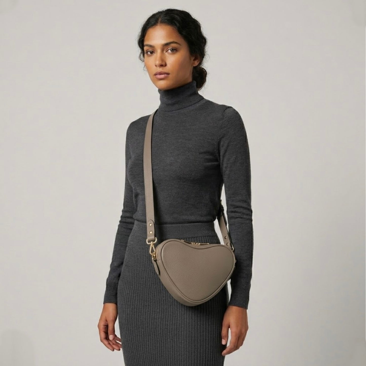 Beige heart-shaped leather crossbody bag by Tikori London, featuring a stylish and compact design. Beautiful dark skinned model wear the bag as a crossbody. She wears grey top and grey tight skirt . Bags long strap and shimmering golden hardware shows in a soft grey background.