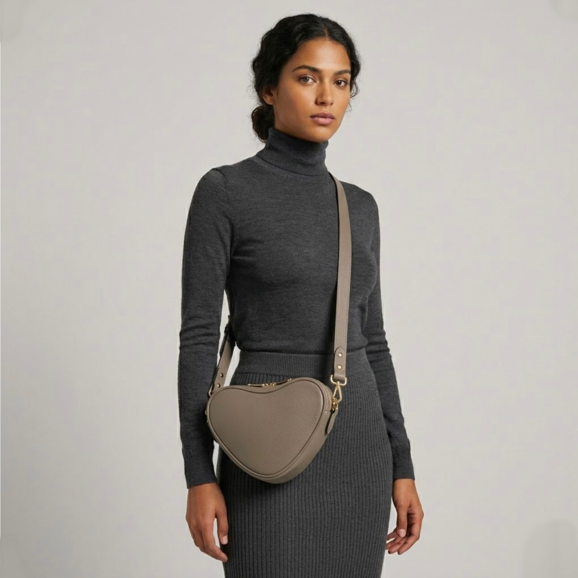 Beige heart-shaped leather crossbody bag by Tikori London, featuring a stylish and compact design. Beautifully dark skinned model wear the bag as a crossbody. She wears grey top and grey tight skirt . Bags long strap and shimmering golden hardware shows in a soft grey background. 