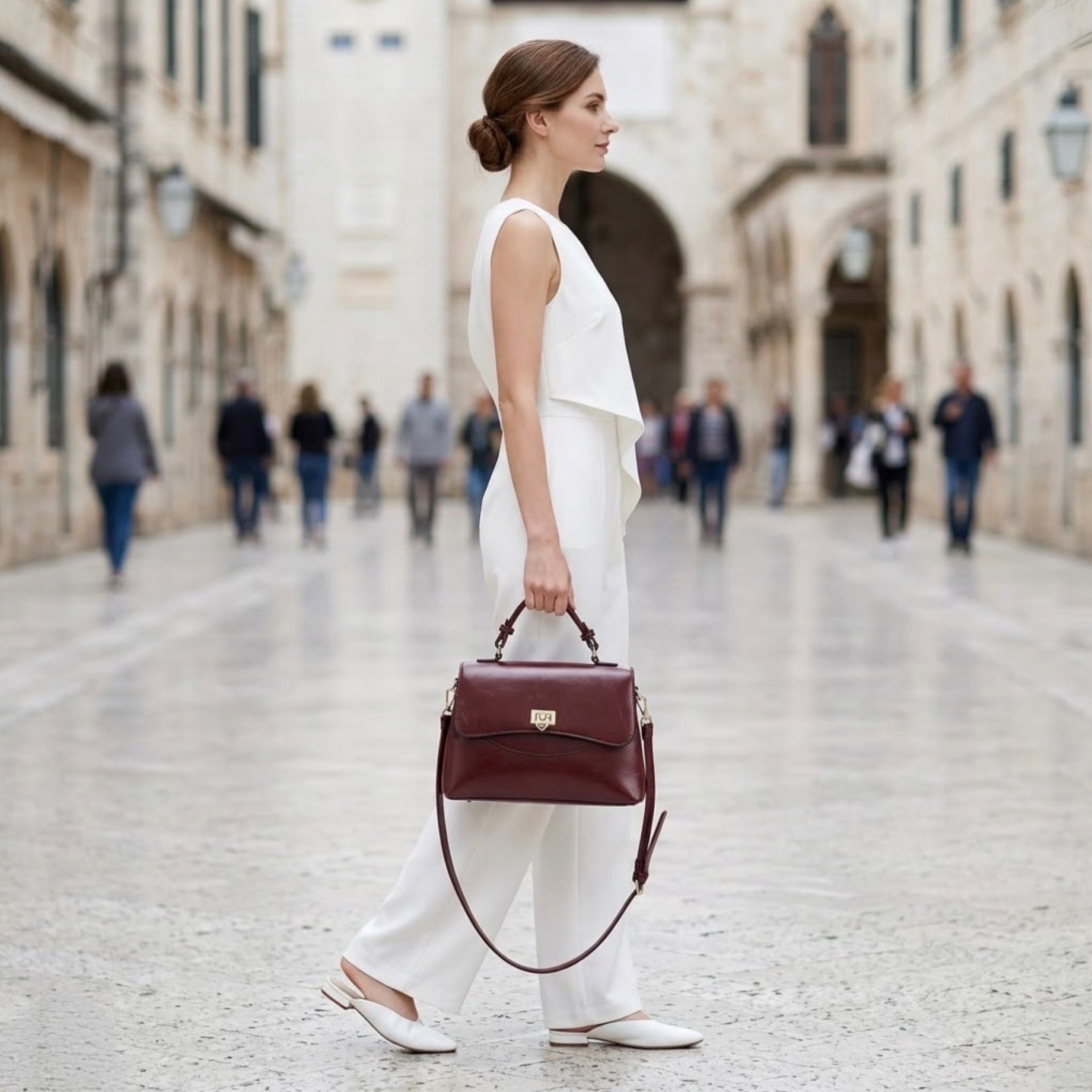 Heritage flap bag maroon by TIKORI LONDON. Defined by a unique braided leather handle that adds a touch of luxury. Features a beautiful, sweeping front flap secured by a polished, shield shaped gold clasp. Model in white top and bottom holding the bag from top handle. Models side profile shows, in side a shopping mall environment.