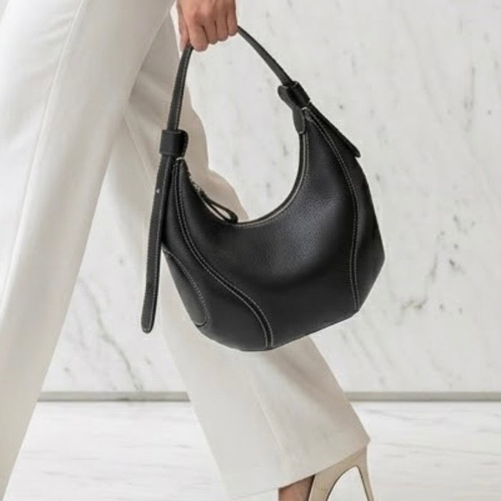 The Ellipse Crescent Hobo Bag black  by TIKORI LONDON, the bag is crafted in premium genuine leather, this piece is defined by its striking artisan contrast stitching that follows the bag's unique ergonomic lines. Women hand seeing carrying the bag in the picture.