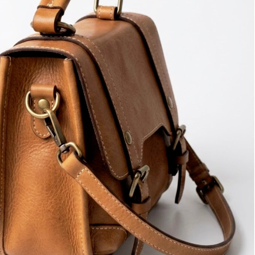 Heritage leather satchel messenger bag with top handle - Warm honey by TIKORI London. Crafted from premium genuine leather, this bag features a structured silhouette that maintains its sophisticated shape whether you’re heading to a boardroom meeting or a weekend brunch.
Side profile of the bag in a white background can be seen in the picture showing brass hardware 