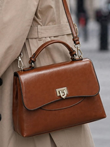 Heritage Flap Bag - Tan  By TIKORI London. Features a beautiful, sweeping front flap secured by a polished, shield shaped gold clasp. Top handle and long strap included. Front of the bag showing on the picture. Middle part of the models body showing wearing the bag as shoulder bag. 