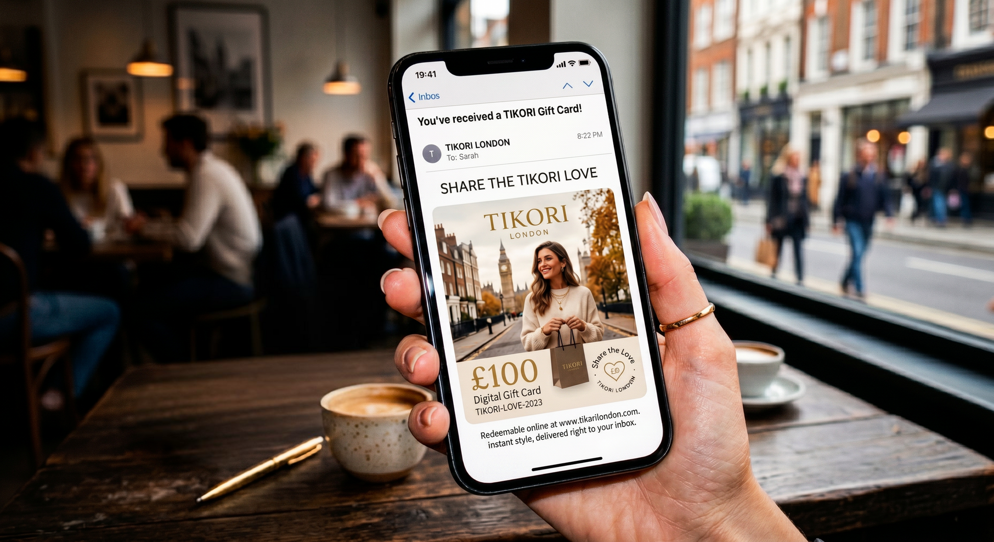 Digital gift card by TIKORI London. Women hands displaying the gift card through the mobile. She’s sitting in a cafe and people are around, showing in the picture. 