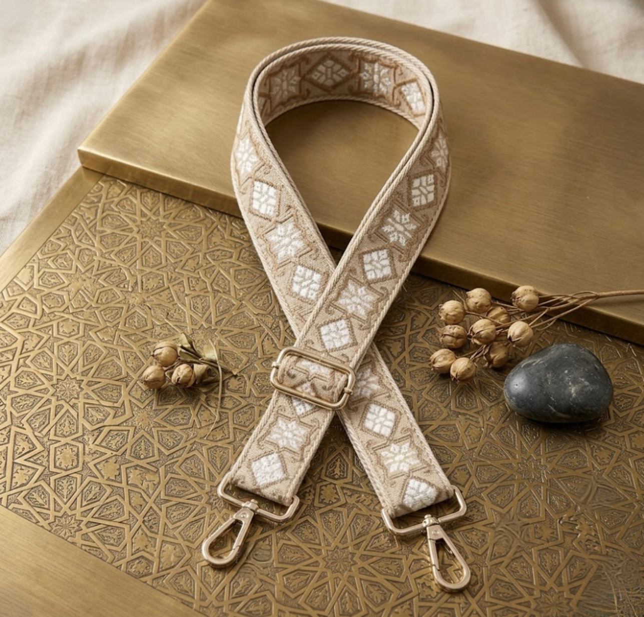 Woven Cross Body Bag Strap - sand Lumi By TIKORI london. Soft geometric patterns with sleek design with golden tone hardware. For instance style upgrade. Strap displays on a golden metal table few dried flower branch and a stone next to it. 