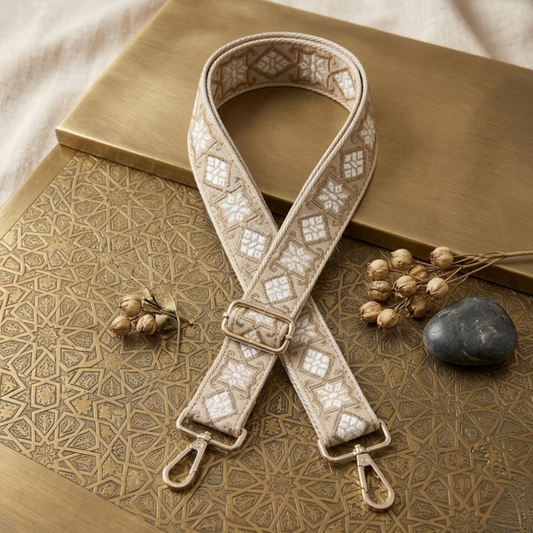Woven Cross Body Bag Strap - sand Lumi By TIKORI london. Soft geometric patterns with sleek design with golden tone hardware. For instance style upgrade. Strap displays on a golden metal table few dried flower branch and a stone next to it. 