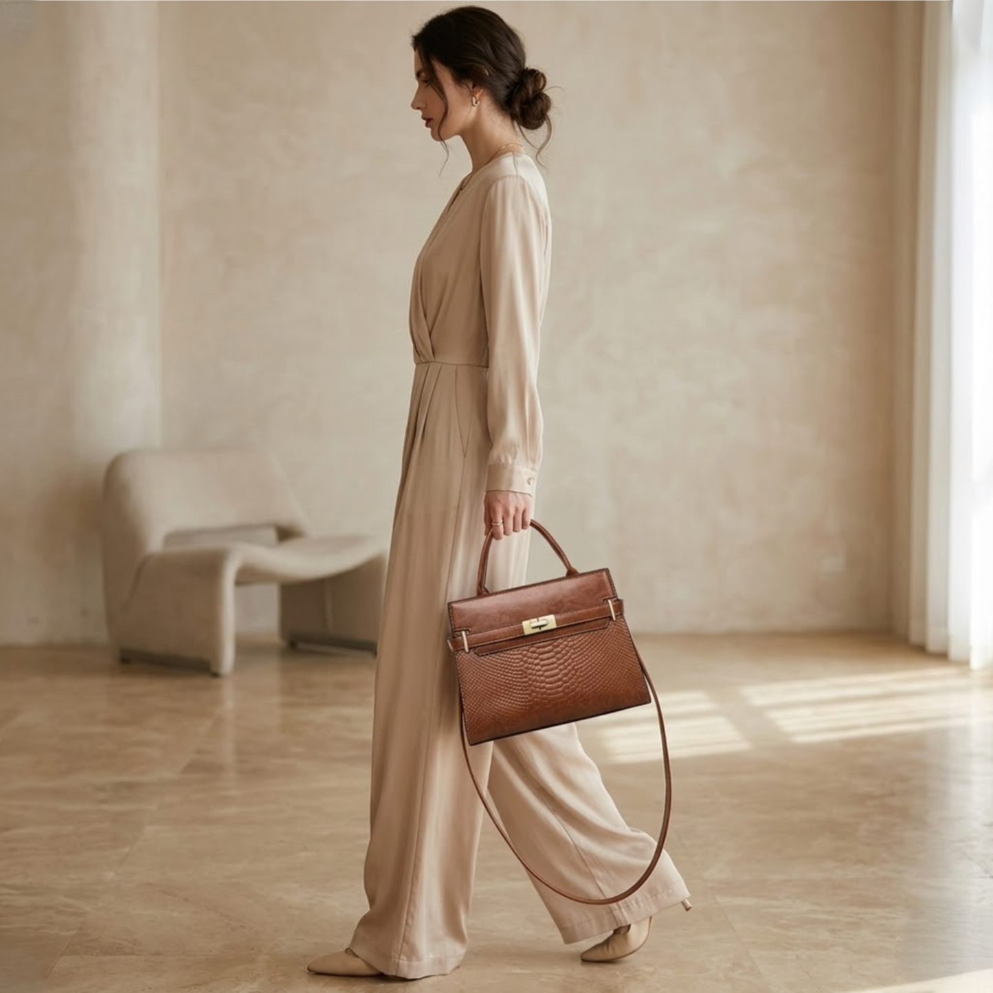 Terra embossed carryall tote tab by TIKORI LONDON. The upper half featured a smooth polished texture, while the lower half features a luxurious, deep embossed reptilian texture for a high contrast in premium high quality Vegan Leather.Beautiful model wearing silky ivory jumpsuit holding the bag in top handle, side profile of the model showing the bag movement. Ivory modern background 