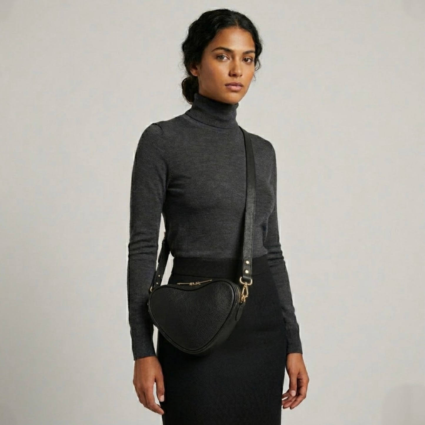 Selene heart crossbody bag by TIKORI London. Genuine premium leather with high quality craftsmanship and exquisite capacity design. Beautiful dark skinned model in black tight shirt and dark grey top wears the crossbody. Long adjustable strap and golden tone hardware shows in soft natural colour background.