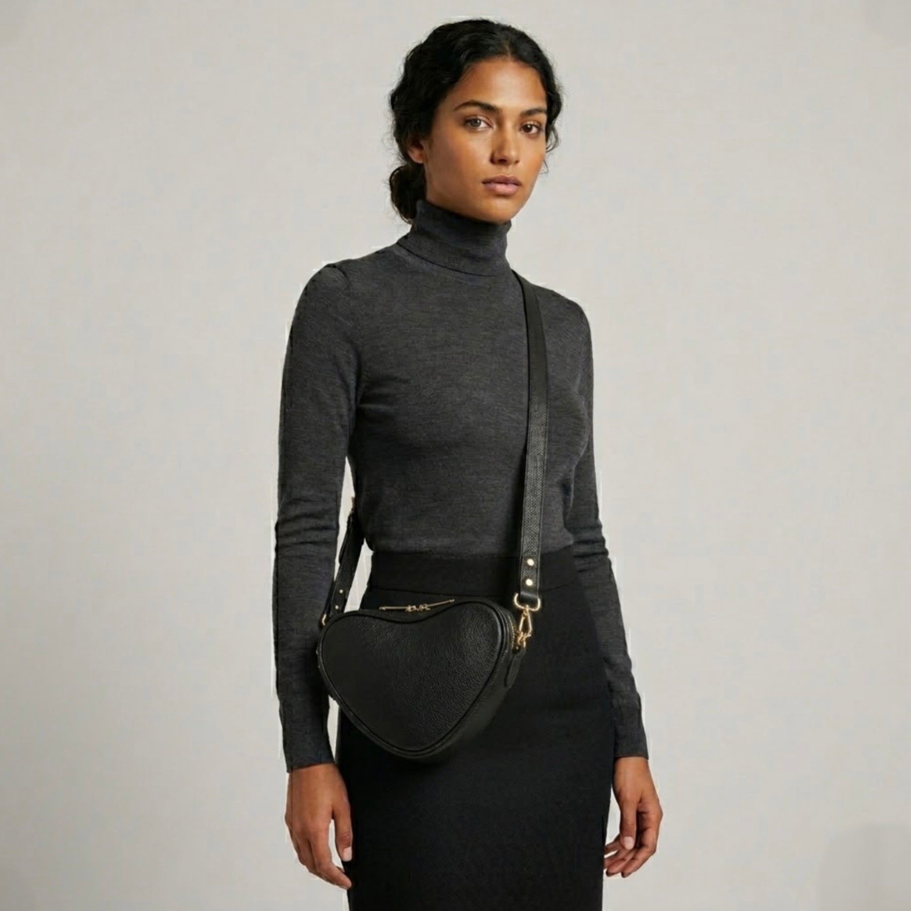 Selene heart crossbody bag by TIKORI London. Genuine premium leather with high quality craftsmanship and exquisite capacity design. Beautiful dark skinned model in black tight shirt and dark grey top wears the crossbody. Long adjustable strap and golden tone hardware shows in soft natural colour background.