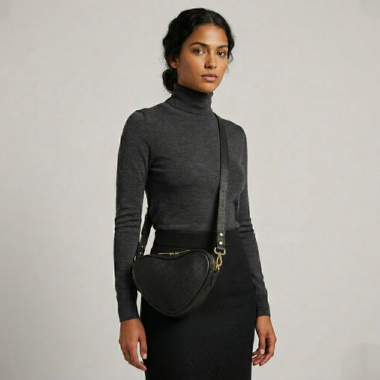 Selene heart crossbody bag by TIKORI London. Genuine premium leather with high quality craftsmanship and exquisite capacity design. Beautiful dark skinned model in black tight shirt and dark grey top wears the crossbody. Long adjustable strap and golden tone hardware shows in soft natural colour background.