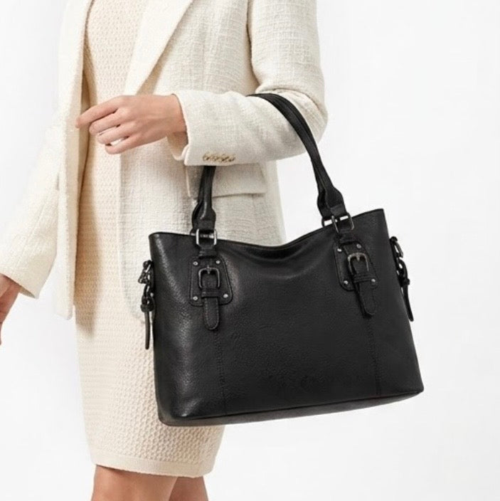 Maven Satchel - Black - by TIKORI LONDON. Constructed from deep-black pebbled Vintage washed Faux Leather with a supple hand-feel, the Marvin is as resilient as it is refined. The gunmetal hardware provides a muted, industrial contrast, ensuring it remains an understated staple rather than a fleeting trend. Front of the bag showing on the picture. With gun metal hardware. Half of the body Model in ivory skirt and blazer showing in the picture, wearing the bag in her hand. In a fully white background.