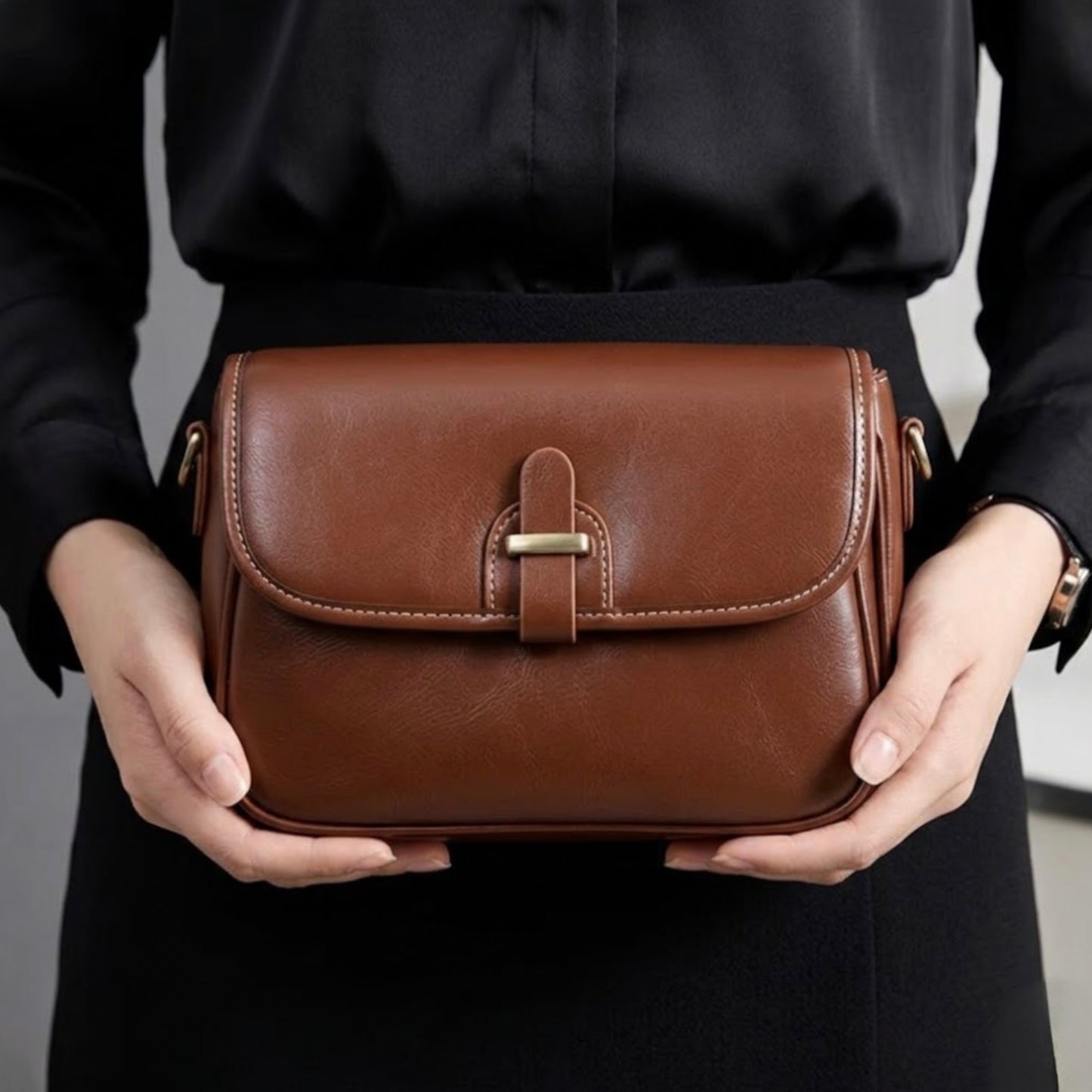 Heritage Cross Body Bag - Mahogany by Tikori London. Bag features a rich, mahogany toned finish that develops a unique patina over time, making it truly yours. Picture shows the front details of the bag  in a white background. Female model wearing a black shirt and skirt holding the bag in both hands in the picture. Models middle part of the body can be seen in the picture.
