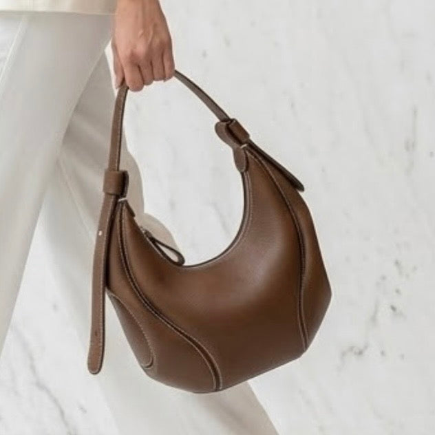 The Ellipse Crescent Hobo Bag brown by TIKORI LONDON, the bag is crafted in premium genuine leather, this piece is defined by its striking artisan contrast stitching that follows the bag's unique ergonomic lines. Women hand seeing carrying the bag in the picture.