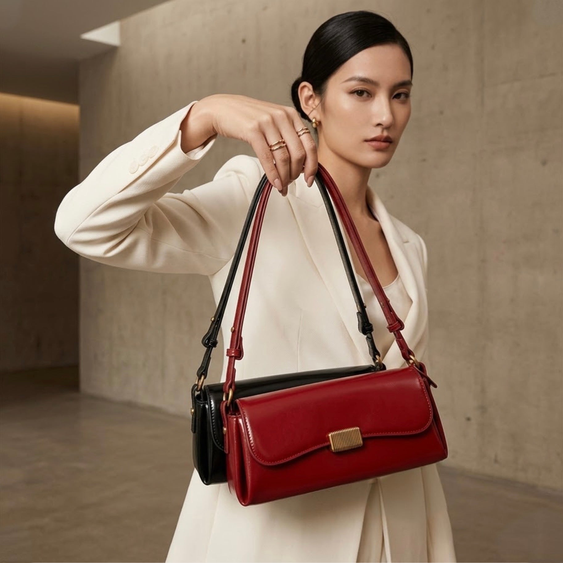 Crimson Muse Shoulder Bag - Black by TIKORI LONDON. Features a structured, elongated "baguette" body with clean, sharp lines and a narrow profile designed to sit snugly under the arm. Picture shows a model wearing a cream blazer , holding both red and black bags high, showing front profile of the bag. 