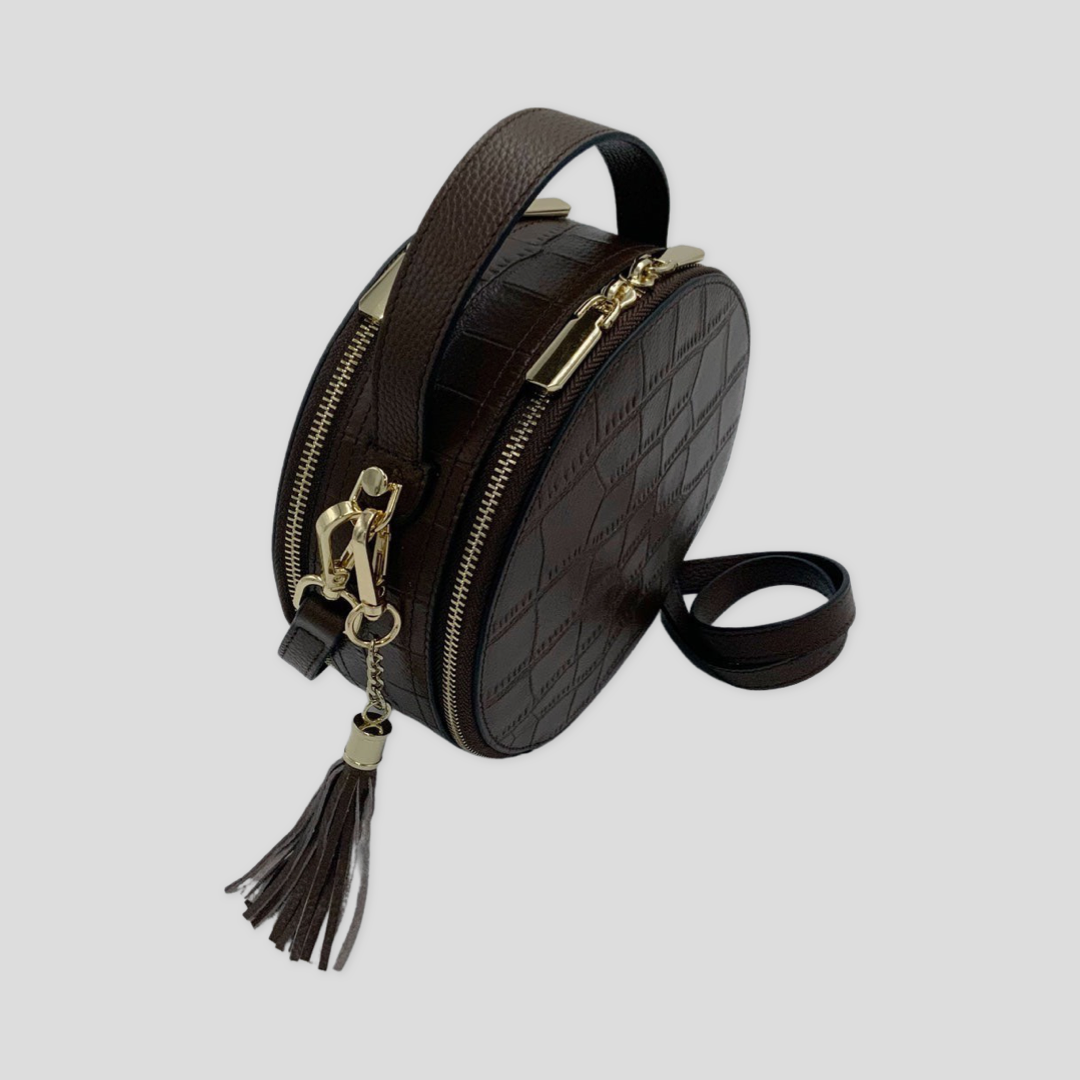 Cleopatra by Tikori London. Dark brown leather circle crossbody bag with dual zip fastenings and finished leather, stylish and compact. Side profile of the bag showing in the picture. Leather strap and dual zipper detail and golden hardware shows in white background.
