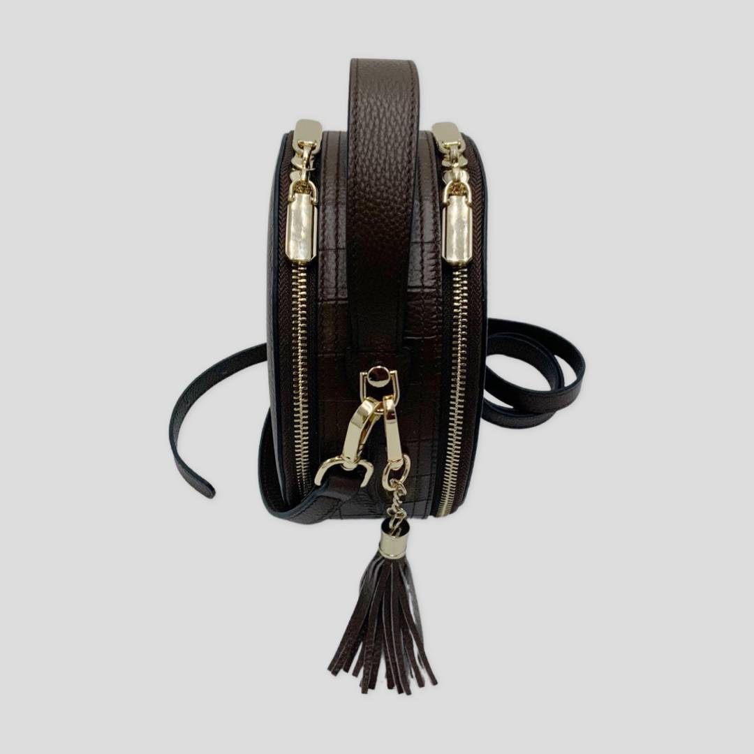 Cleopatra by Tikori London. Dark brown leather circle crossbody bag with dual zip fastenings and finished leather, stylish and compact. Side profile of the bag showing golden hardware zipper detail strap and tussle in white background 