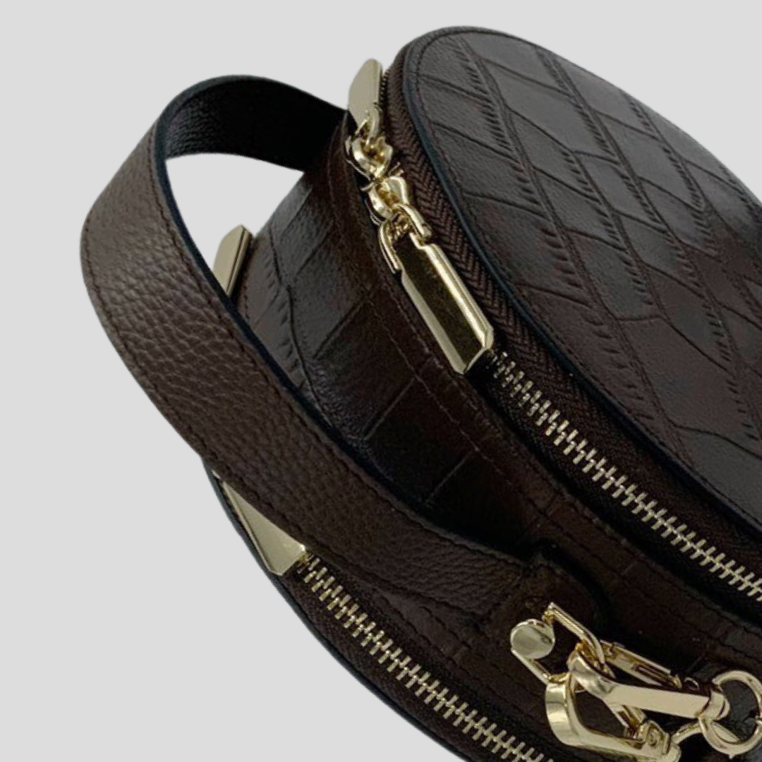 Cleopatra by TIKORI london. Dark brown leather circle crossbody bag with dual zip fastening. featuring a sleek finished leather design. Close up detail of the handle showing in the picture, with golden hardware.