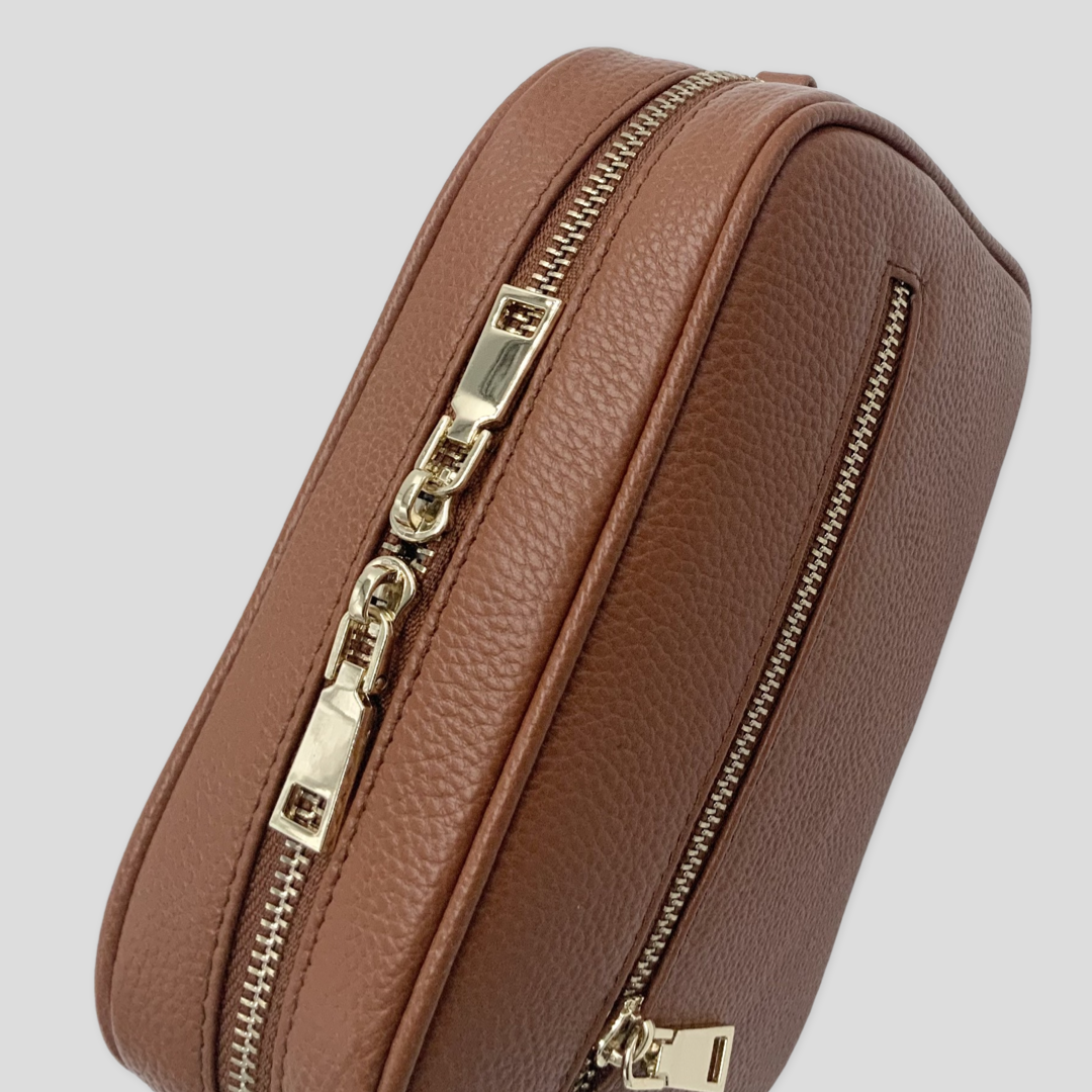 Classic elegance. TIKORI London classic pebbled leather crossbody bag in warm tan with adjustable strap and sleek design. Front close profile of the bag showing in the picture. Golden zipper and front pocket is highlighted. Close up detail of the golden hardware shows in white background .