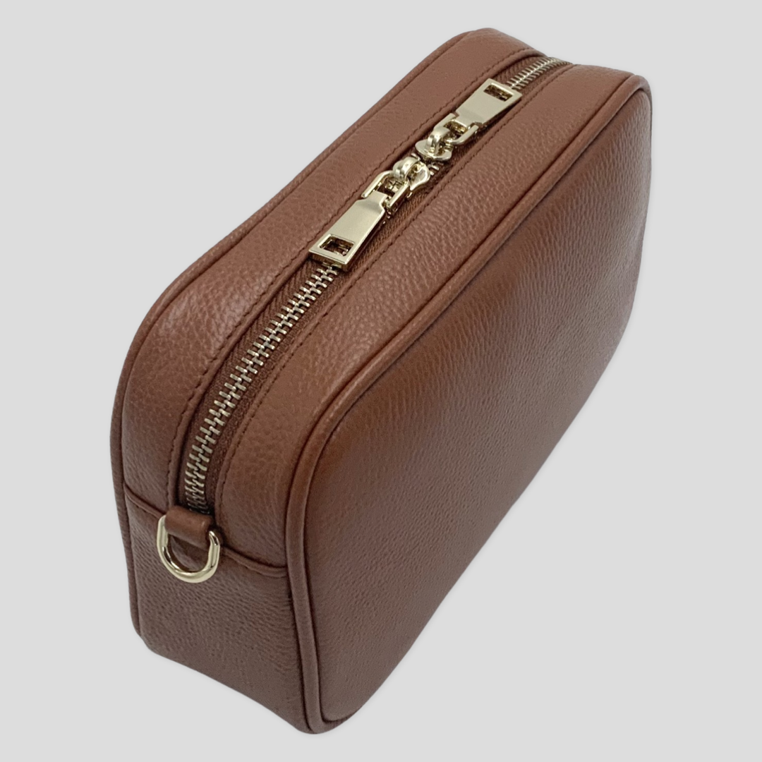 Classic elegance. TIKORI London classic pebbled leather crossbody bag in warm tan with adjustable strap and sleek design. Back profile of the bag showing in the picture. Close up detail of the golden hardware shows in white background 