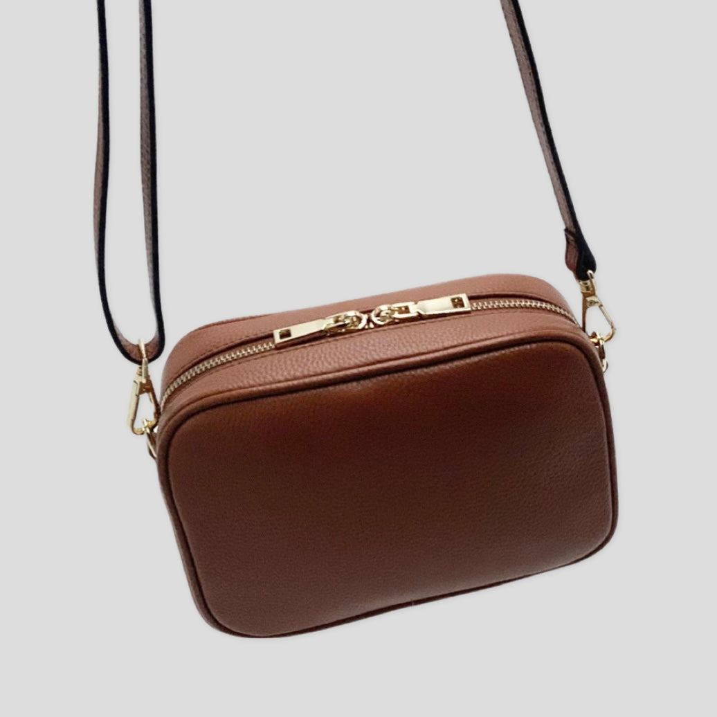 Classic elegance. TIKORI London classic pebbled leather crossbody bag in warm tan with adjustable strap and sleek design. Back  profile of the bag showing in the picture in hanging motion. Long leather strap shows and Close up detail of the golden hardware shows in white background 