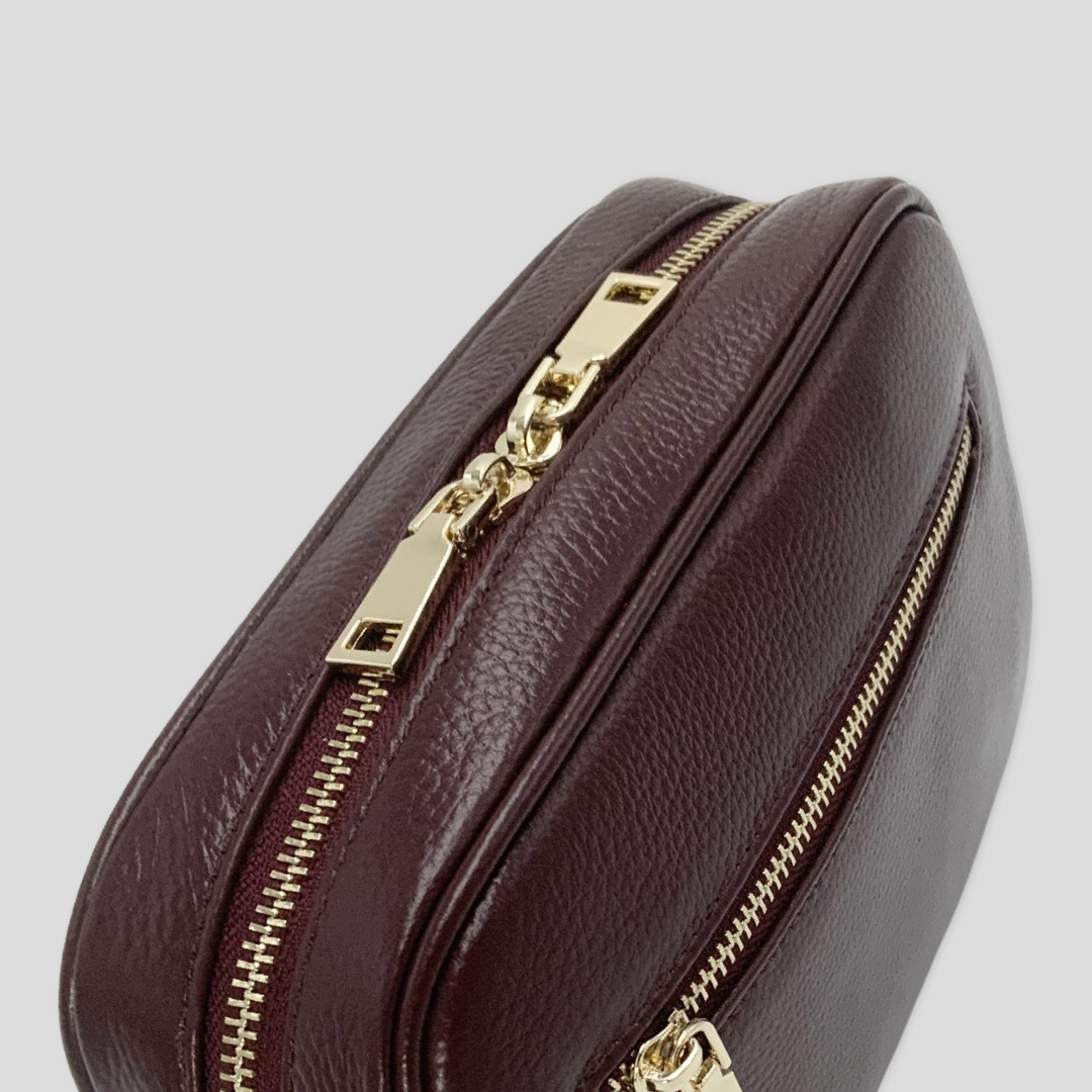 Classic elegance with practicality and versatility in mind. TIKORI classic maroon by TIKORI london. Genuine Leather crossbody bag. Close profile of top of the bag showing with zipper detail with golden tone hardware and front pocket in white background. 