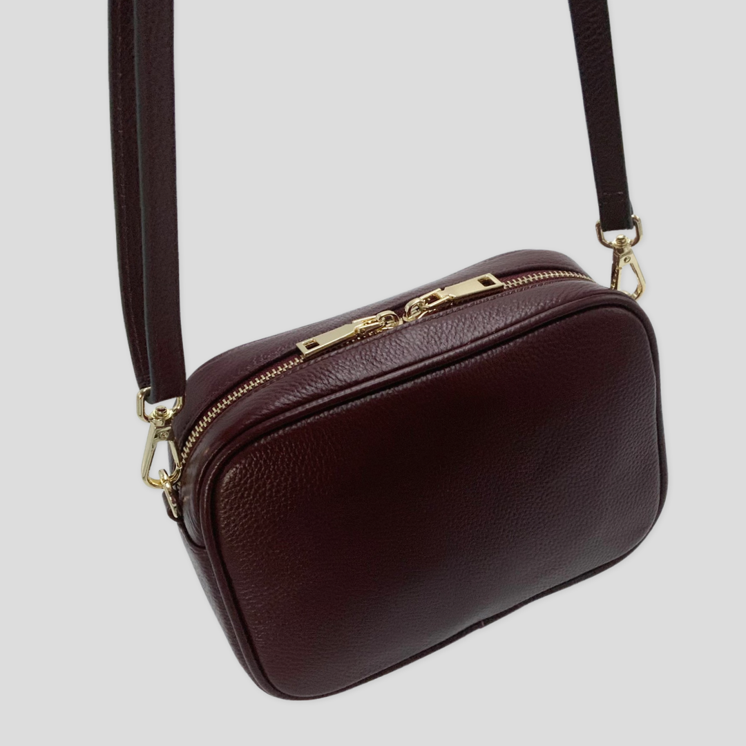 Classic elegance with practicality and versatility in mind. TIKORI classic maroon by TIKORI london. Genuine Leather crossbody bag. Back profile of the bag showing with golden tone hardware in hanging motion . Long strap shows in white background. 