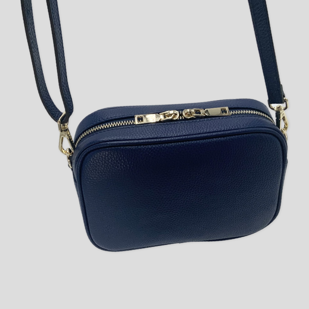 Classic elegance with practicality and versatility in mind. TIKORI classic Navy by TIKORI london. Genuine Leather cross body bag. Back  profile of the bag showing with golden tone hardware in hanging motion. Top zipper with golden hardware shows in Fully white background.