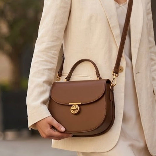 Luna saddle bag - Brown TIKORI LONDON. Body of a Model wearing the bag as crossbody. She wears silky cream dress and scream blazer. Front profile of the bag showing long strap and t bar lock hardware.