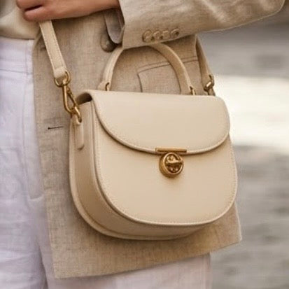 Luna Saddle Bag - Ivory by TIKORI LONDON. The centrepiece is a custom engineered gold T-bar turn lock, providing a secure closure and a sophisticated focal point. Hip area of a model showing wearing the bag as a crossbody in Mediterranean background. White cotton trousers and cream blazer can be seen. Bag featuring Golden tone hardware and long strap. Front profile of the bag in the picture.