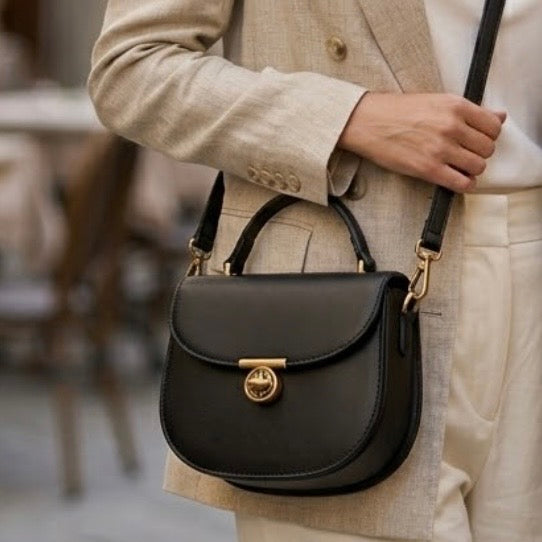 Luna saddle bag - Black - TIKORI LONDON. Model wearing the bag as crossbody. Close up detail of a model wearing the bag as crossbody . She wears cream trousers and cream blazer. Front profile of the bag showing long strap and t bar lock hardware.