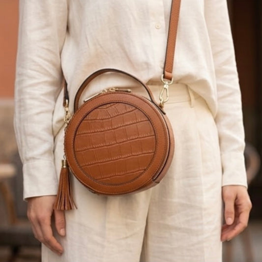 Cleopatra by TIKORI London. warm tan leather circle crossbody bag with dual zip fastening and finished leather details. Model in ivory cloths wearing the bag and front profile of the bag, golden hardware and long strap showing in the picture.