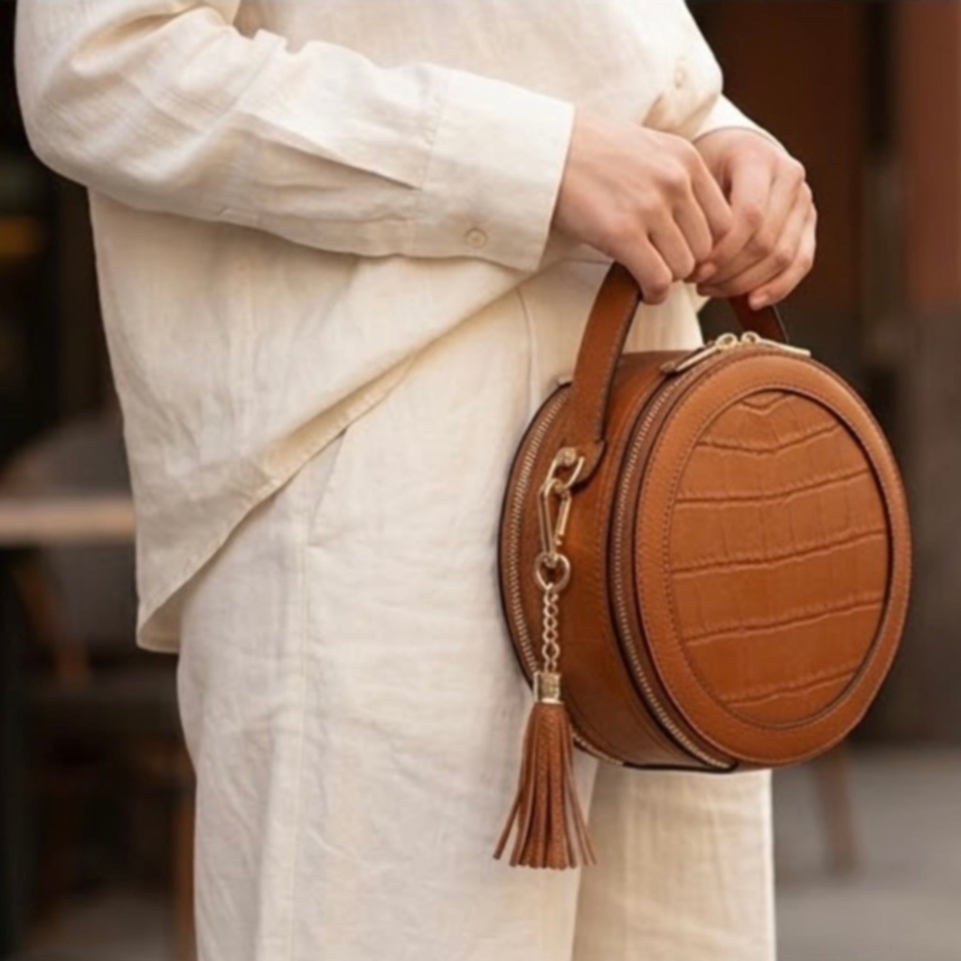 Cleopatra by TIKORI London. warm tan leather circle crossbody bag with dual zip fastening and finished leather details. Model in Ivory cloths holding the bag from top handle showing in the picture. Golden hardware and hanging tussle shows in the picture.