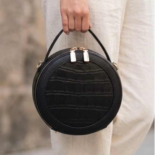 TIKORI LONDON black finished leather circle crossbody bag with dual zip fastenings, featuring a sleek, rounded design. Body of a model in ivory cloths carrying the bag from top handle. Front profile of the bag with golden hardware shows in the picture 