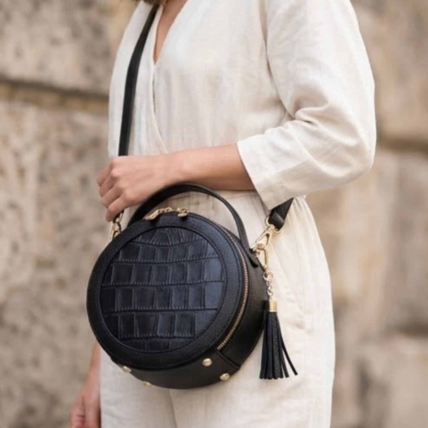 TIKORI LONDON black finished leather circle crossbody bag with dual zip fastenings, featuring a sleek, rounded design. Body of a model in ivory cloths wearing the bag as cross body. Front profile of the bag with golden hardware and tussle detail shows in the picture 
