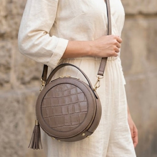 Cleopatra Mink by Tikora’s London. leather circle crossbody bag collection with dual zippers, crafted in leather with sleek, modern design. Model in ivory cloth wearing the bag in the picture. Front profile of the bag showing with golden hardware and hanging tussle. 
