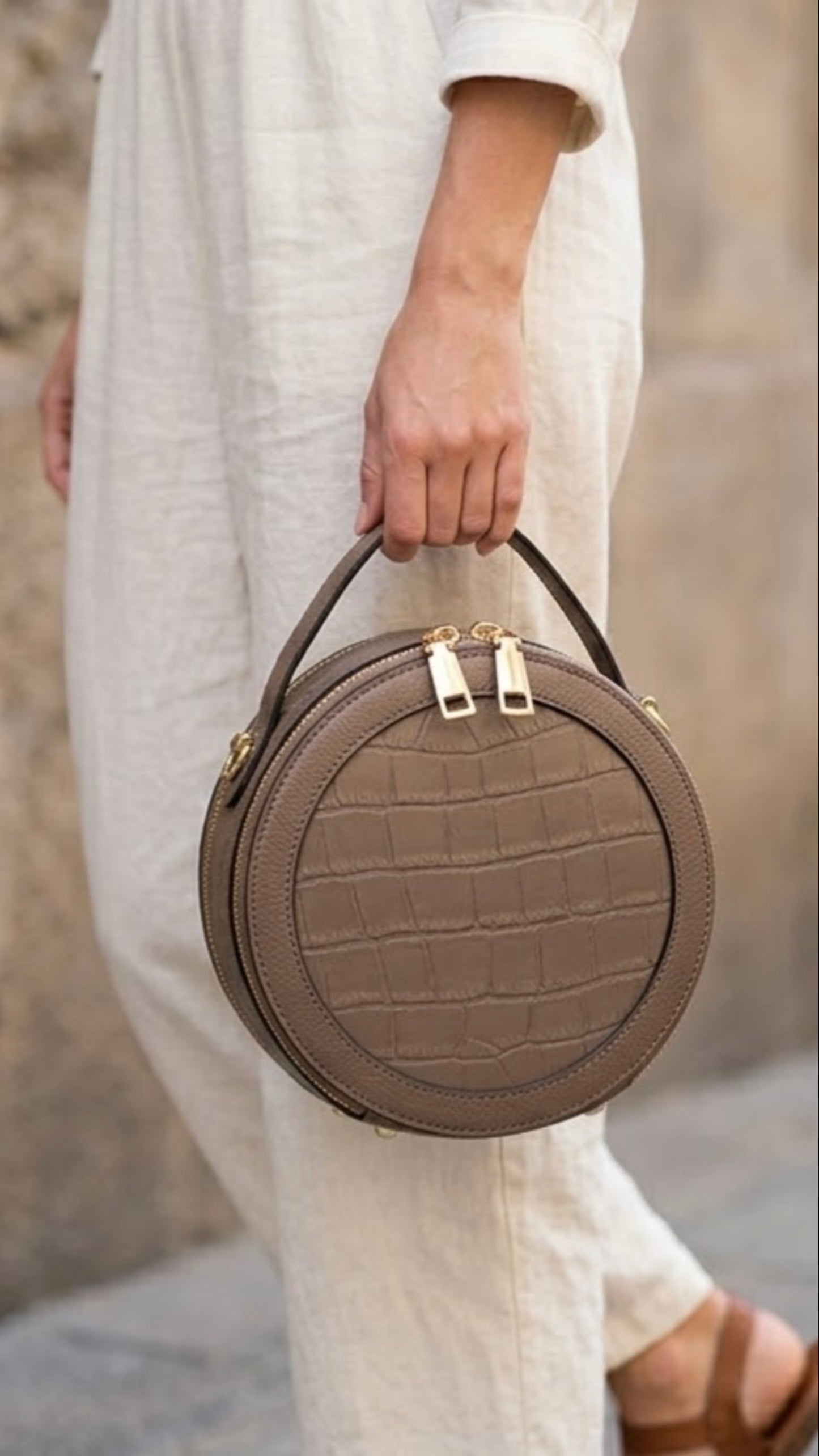 Cleopatra Mink by Tikora’s London. leather circle crossbody bag collection with dual zippers, crafted in leather with sleek, modern design. Model in ivory cloths holding the bag from top handle. Front profile of the bag showing with golden hardware.