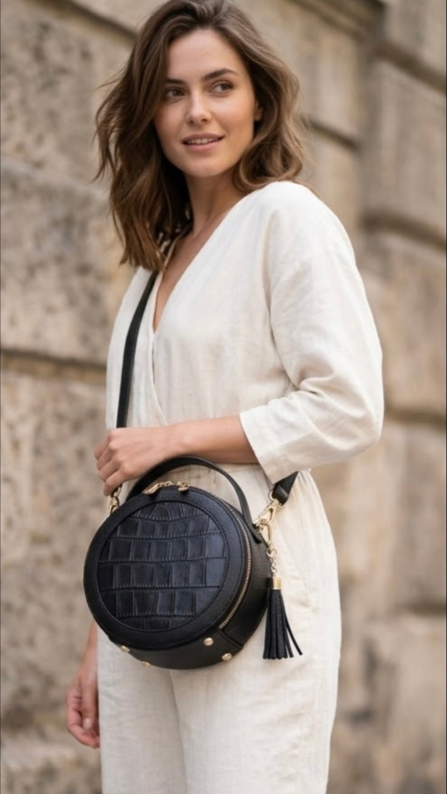 TIKORI LONDON black finished leather circle crossbody bag with dual zip fastenings, featuring a sleek, rounded design. Model in ivory cloths wearing the bag as cross body. Front profile of the bag with golden hardware and tussle detail shows in the picture 