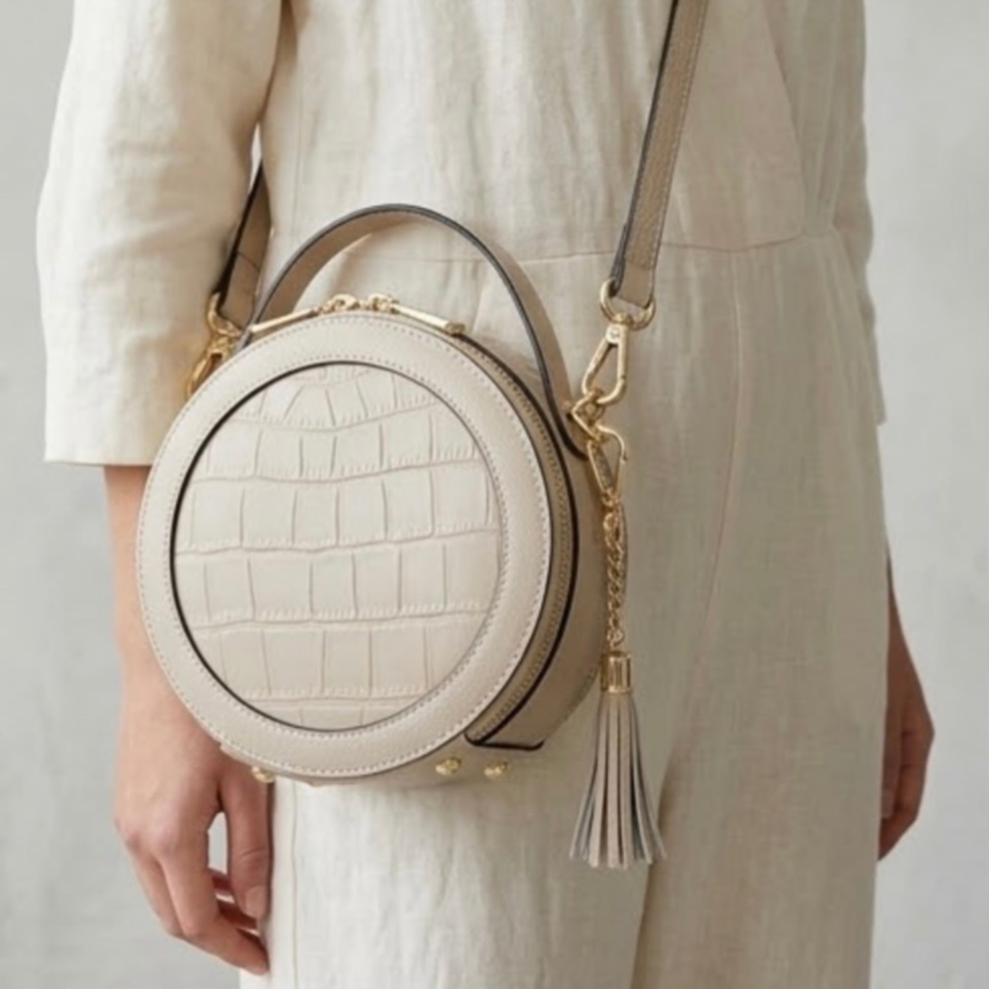 Cleopatra by TIKORI London. Ivory leather circle crossbody bag with dual zip fastening and finished leather details. Body of a Model in ivory cloths wearing the bag as cross body. Front profile of the bag, with golden hardware and tussle on the side shows in the picture.