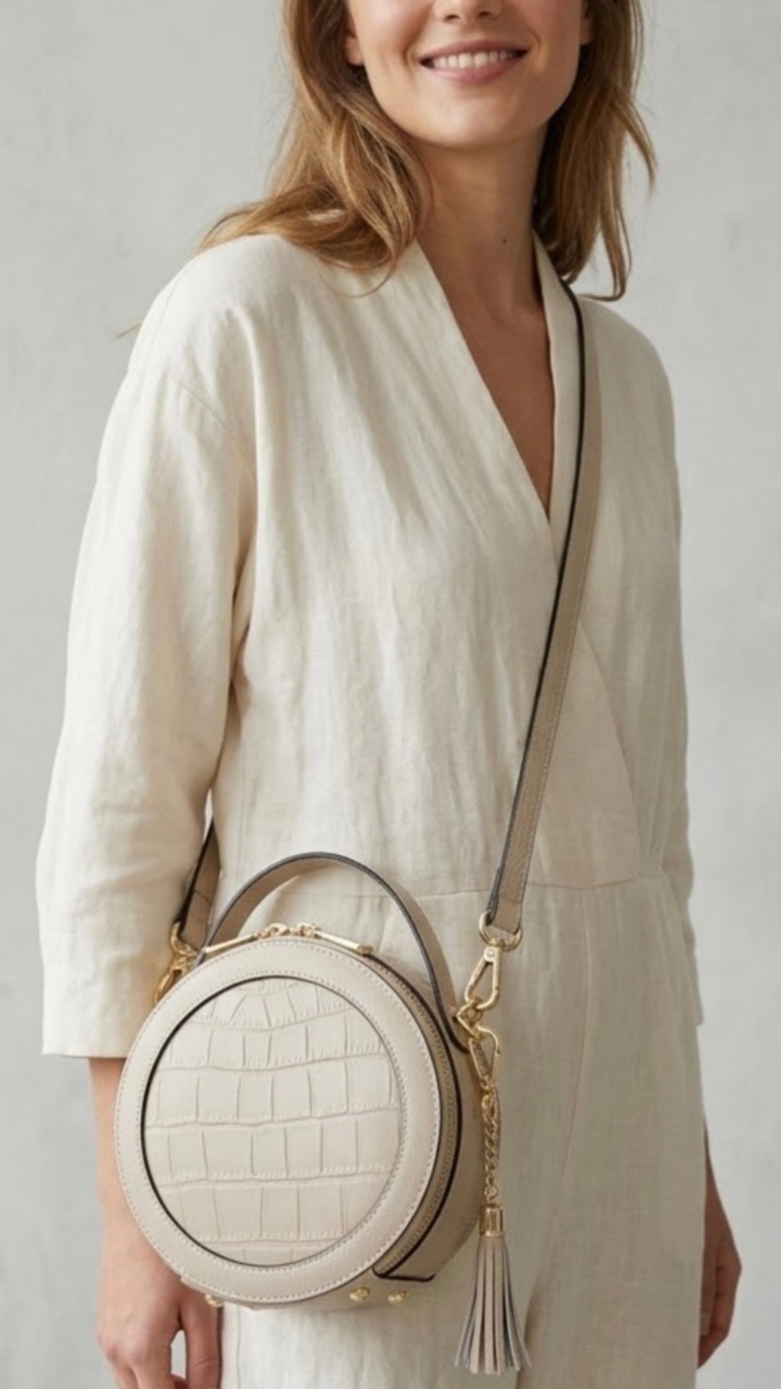 Cleopatra by TIKORI London. Ivory leather circle crossbody bag with dual zip fastening and finished leather details. Smiling Model face. She is  in ivory cloths wearing the bag as cross body. Front profile of the bag, with golden hardware and tussle on the side shows in the picture.