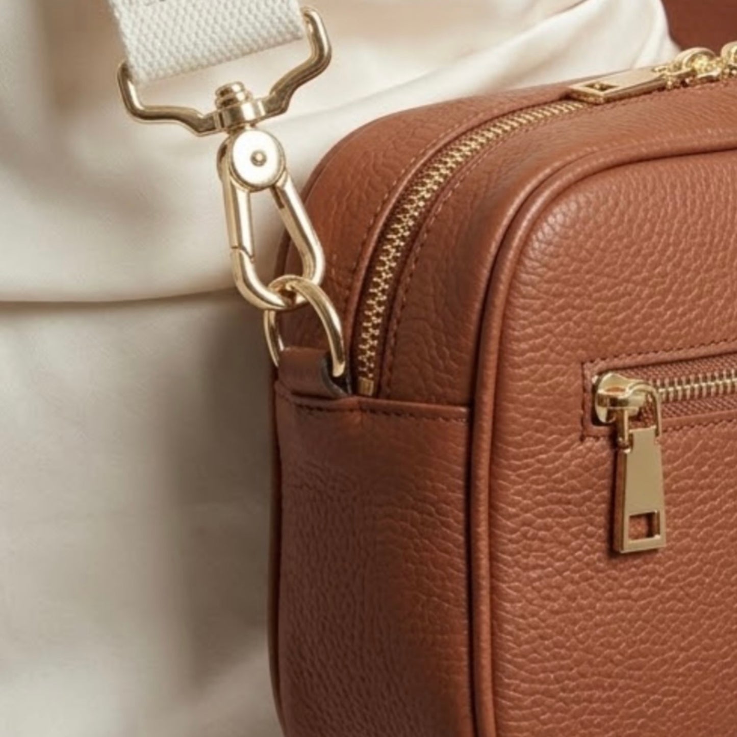 Classic elegance. TIKORI London classic pebbled leather crossbody bag in warm tan with adjustable strap and sleek design. Model wears the bag. Front profile of the bag shows, close up detail of the golden hardware in white background. front profile of the bag with golden zipper detail. 