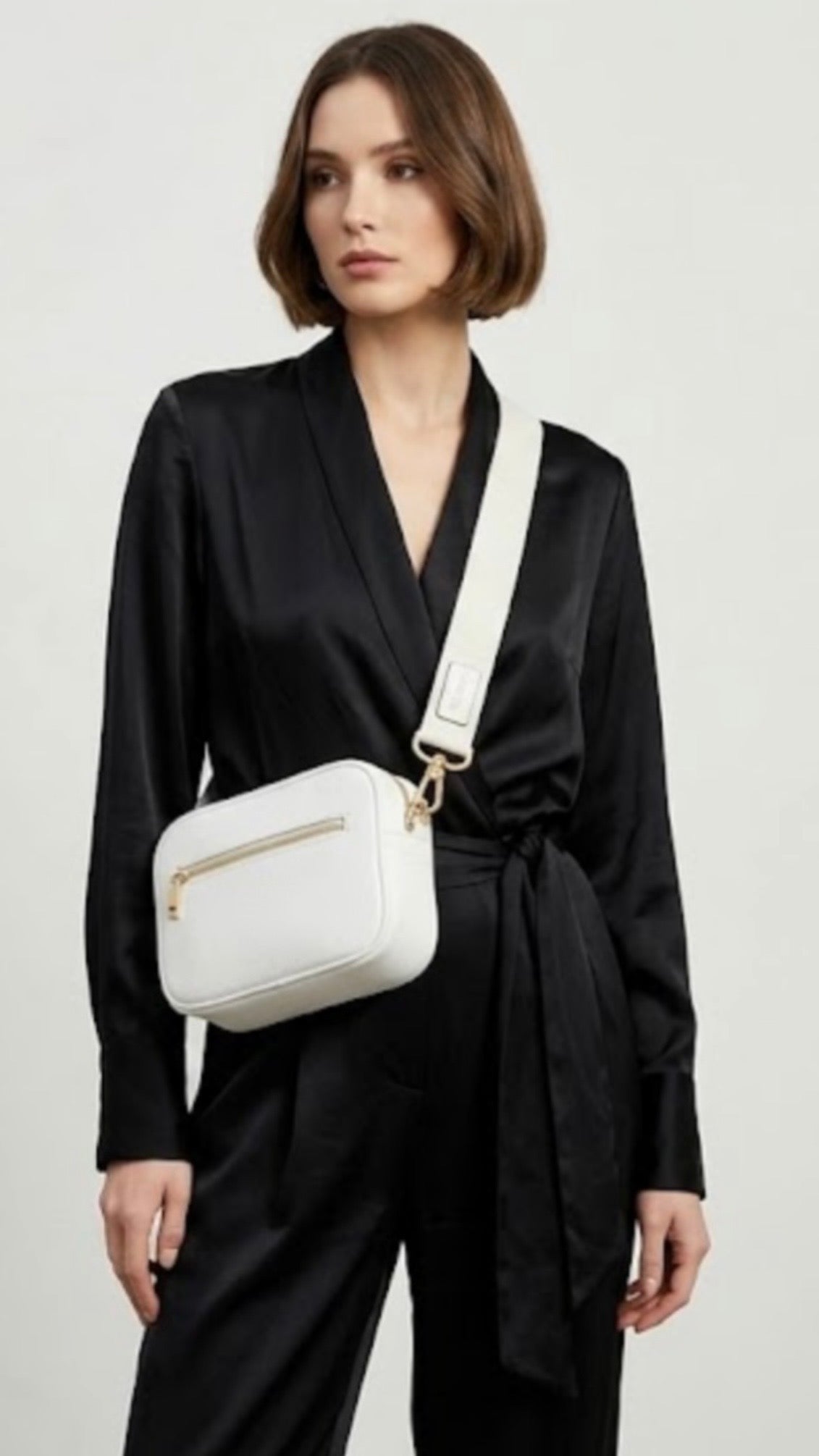 Classic pebbled leather cross body bag - White - TIKORI LONDON. Model in black silk cloths wears the cross body. Front  profile of the bag showing the zipper details and gold hardware in white background.
