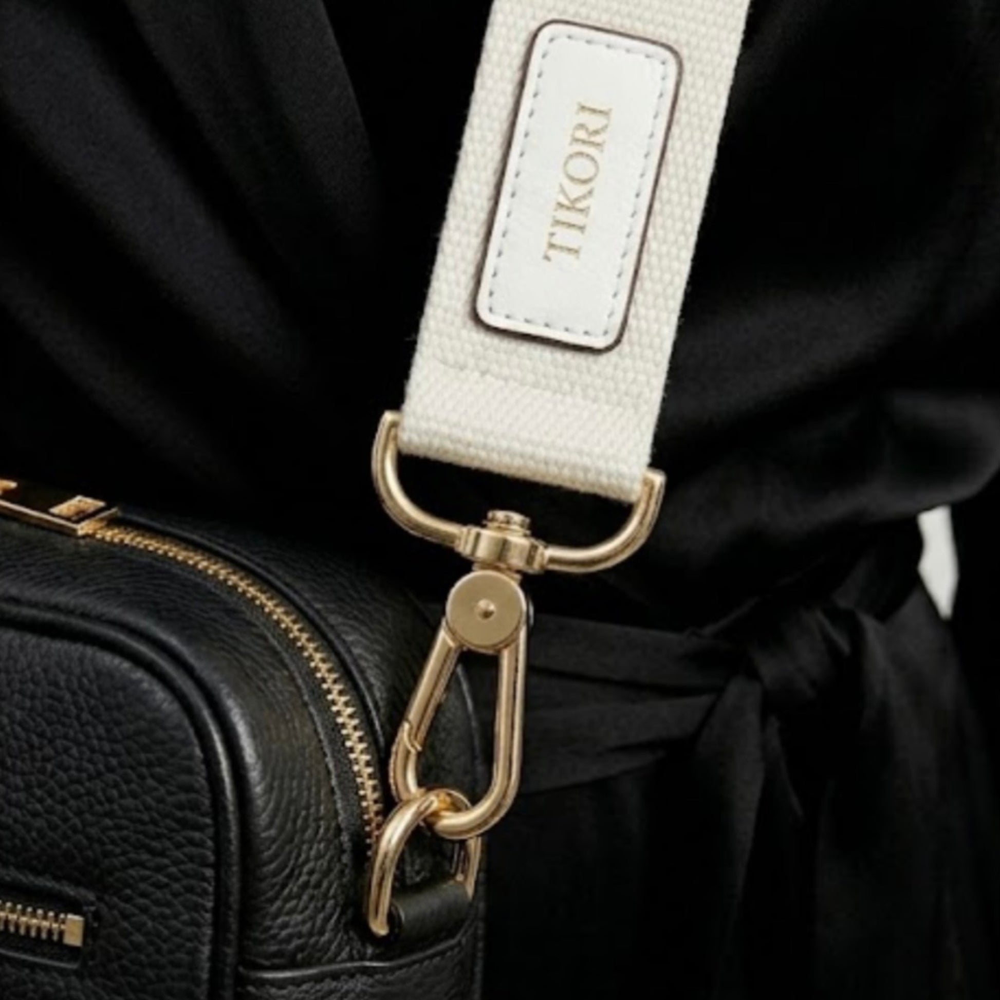 Classic elegance with practicality and versatility in mind. TIKORI classic Black by TIKORI london. Model in black cloths, wears the crossbody. Close up detail of the canvas strap and golden tone hardware shows in the picture with TIKORI logo on the canvas strap. 